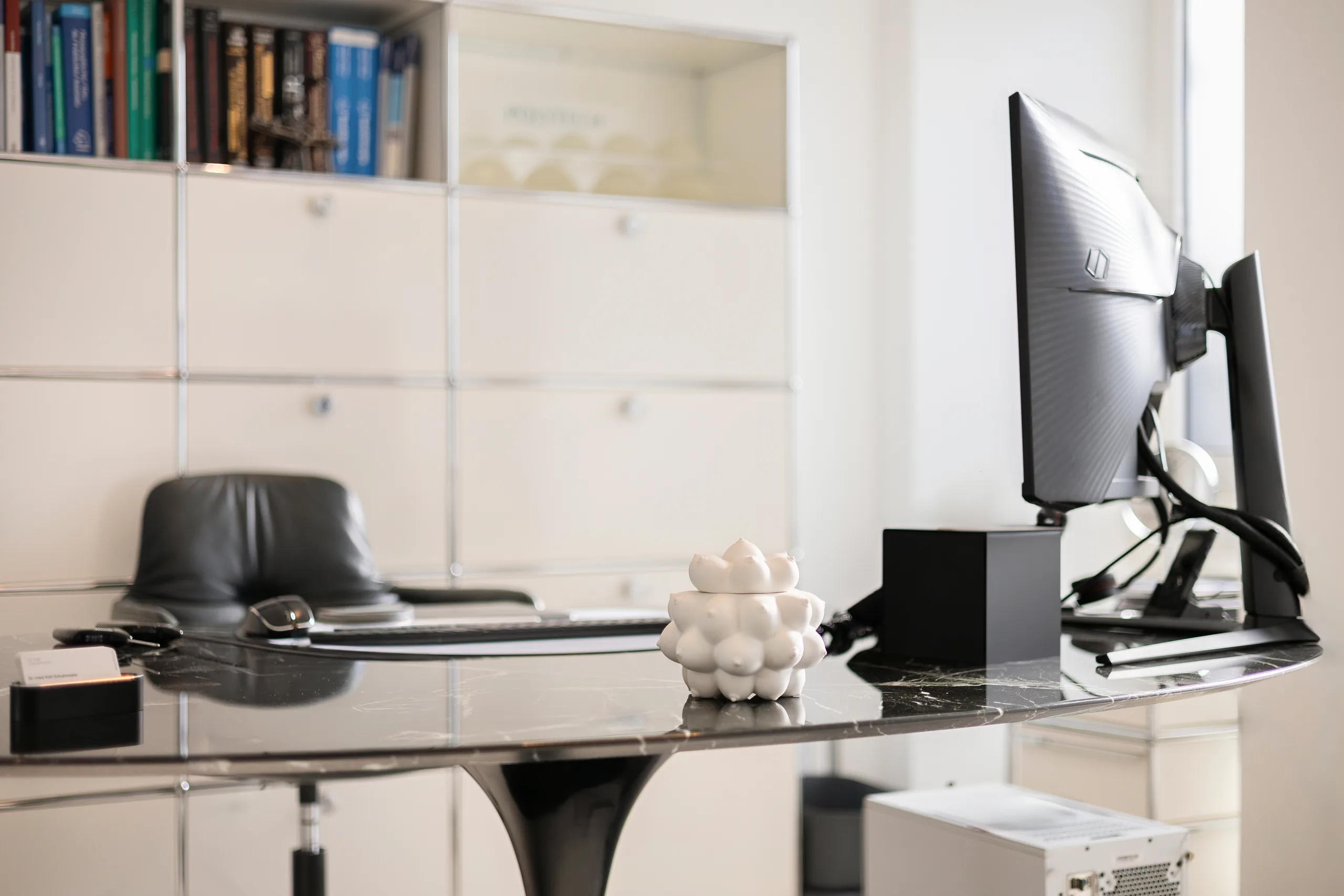 Consultation room at the artethic® practice in Düsseldorf with a black glass desk, a monitor and a white sculpture.