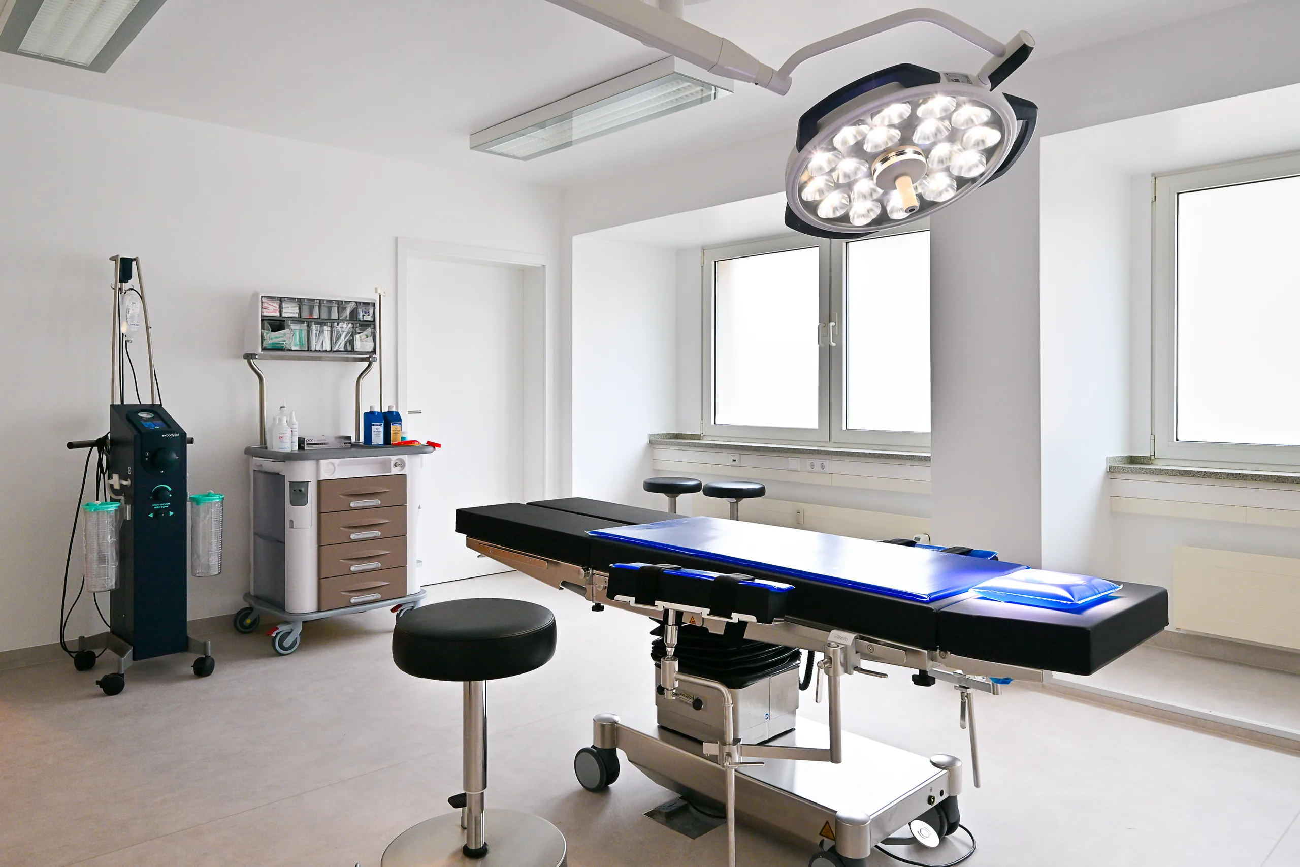 Eingriffsraum der Praxis artethic® in Düsseldorf mit OP-Leuchte, OP-Tisch und medizinischen Geräten am Fenster.